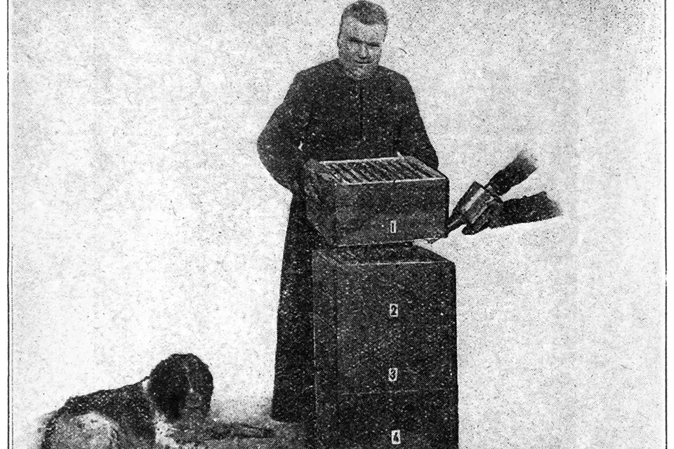 Photographie ancienne en noir et blanc : l'abbé Warré en soutane, debout à côté d'une ruche faite de caisses empilées, tenant une caisse à barrettes, un chien couché à ses pieds